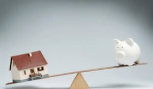 a house and piggy bank sitting on opposite sides of a see-saw with the house weighing down the piggy bank