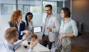 a group of five people in an office setting all looking at a chart on a tablet held by a man