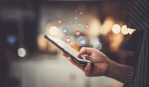 hand holding a cell phone with computer-generated thumbs up like icons and heart icons floating above